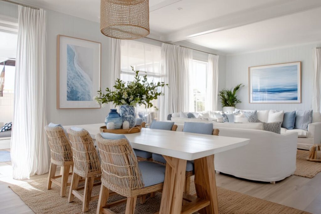 Helles Wohnzimmer und Esszimmer im maritimen Einrichtungsstil mit weißen Möbeln, blauen Akzenten, Rattan-Stühlen und Wandbildern mit Meeresmotiven